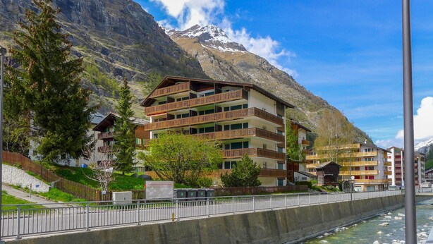 Ein mehrstöckiges Hotel mit Balkonen, im Vordergrund ein Fluss und im Hintergrund Berge.