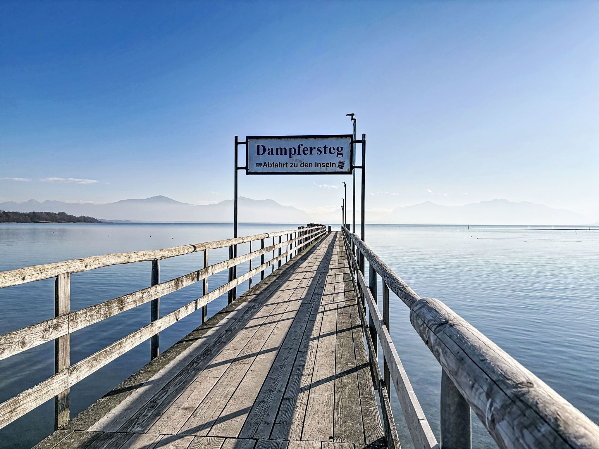 A wooden pier leading to a sign reading "Dampersteg" with a view of calm water and distant mountains.