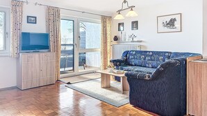 Een moderne woonkamer met een blauwe bank, een houten salontafel, een tv op een kast en een groot raam met gordijnen.