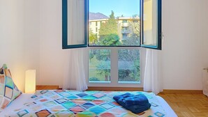 Ein Schlafzimmer mit einem großen Fenster, einem Bett mit farbenfroher Decke und Blick auf Gebäude und Bäume.