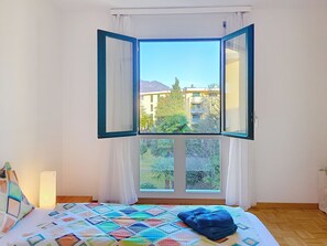 A bedroom with a large window, a bed with a colorful quilt, and a view of buildings and trees outside. - Apt. Hermosa by Interhome (Locarno)