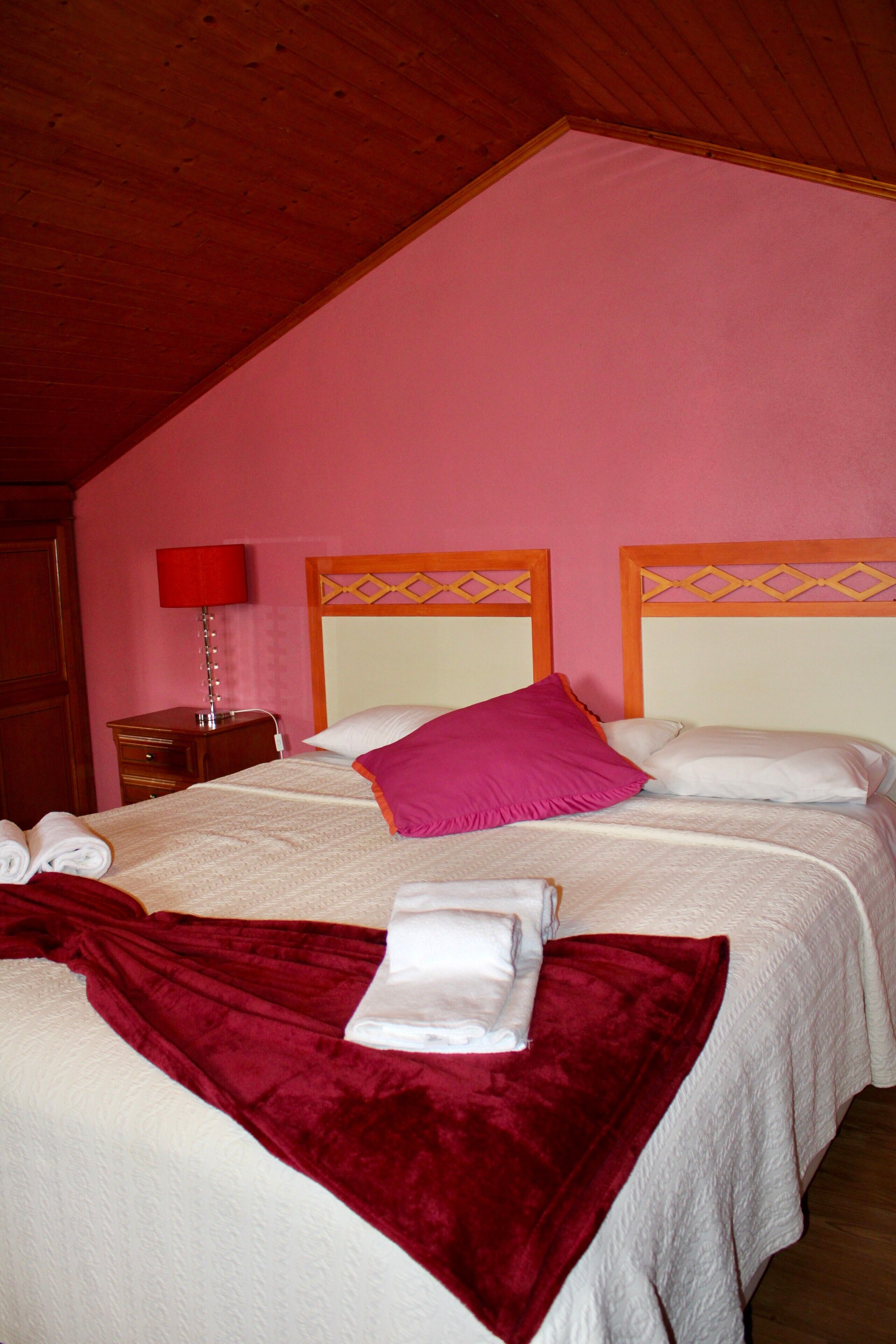 A hotel room with a wooden ceiling, a bed with white bedding and a red throw, a nightstand with a red lamp, and a pink wall.
