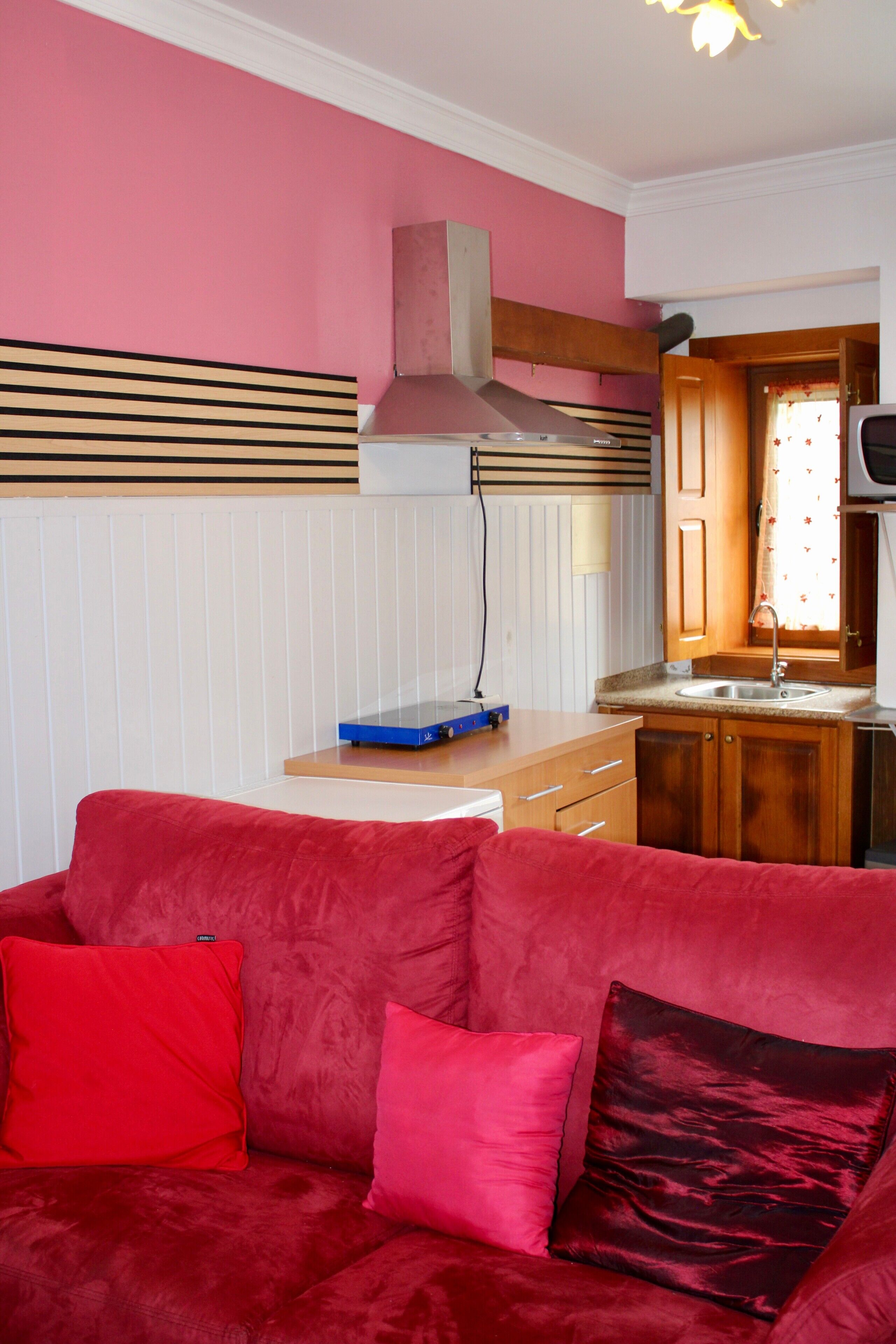 Un espace de vie compact comprenant un coin cuisine, un canapé rouge avec des coussins et un téléviseur.
