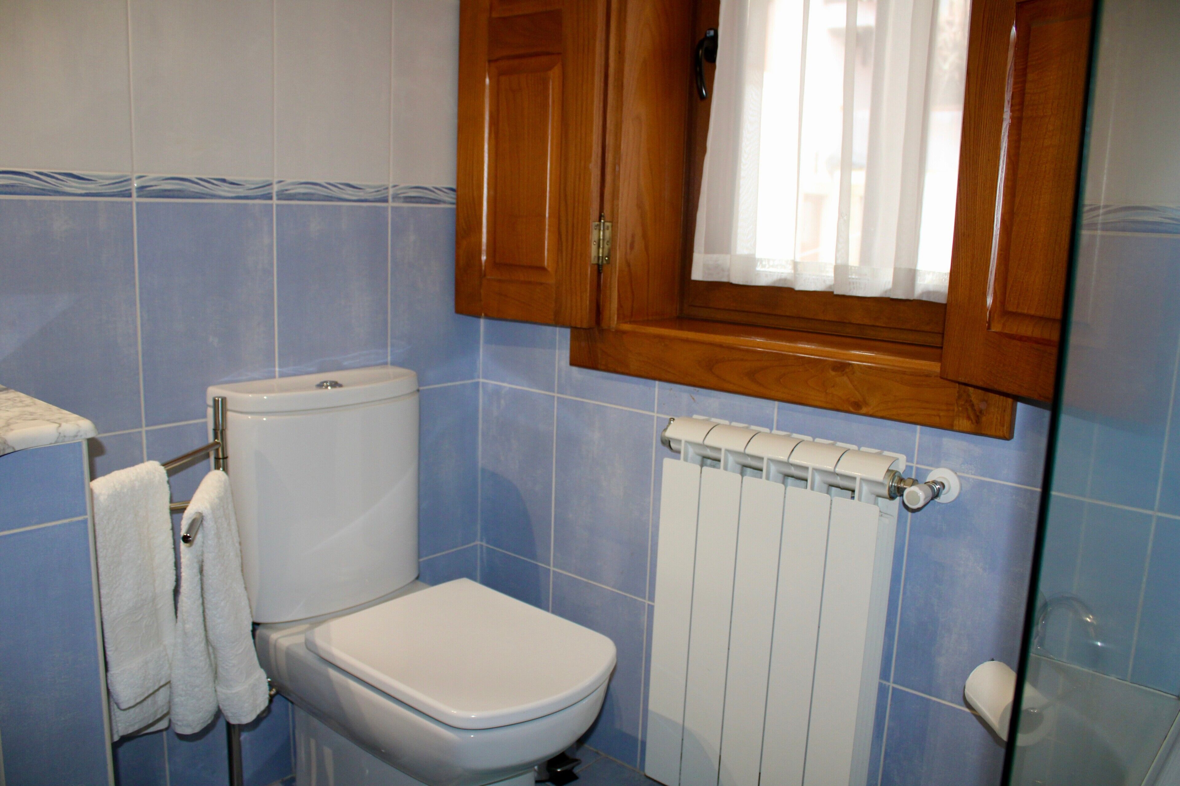 A bathroom with a toilet, a towel rack with towels, a wooden cabinet, and a window with a curtain.