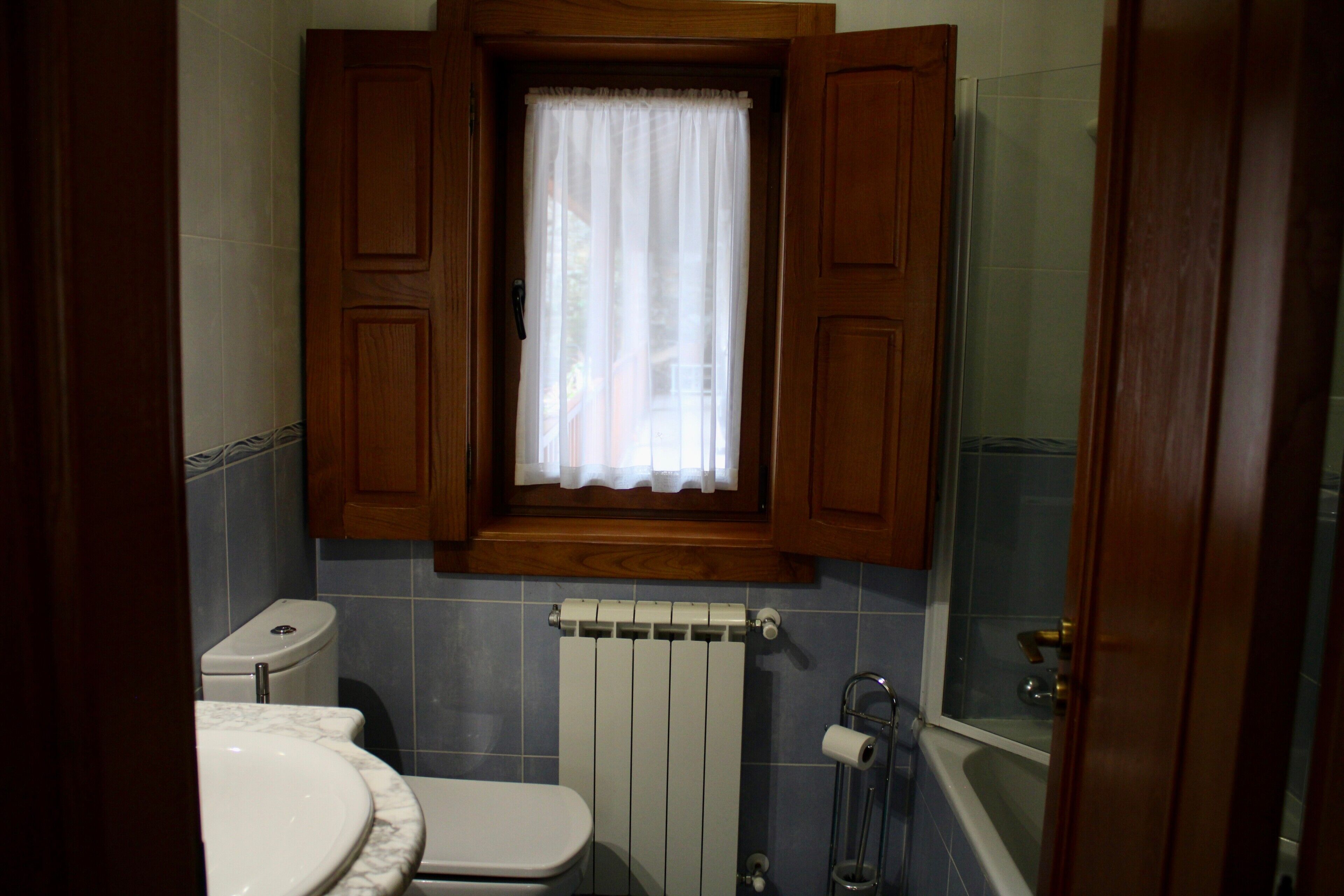 A bathroom with a toilet, a sink, a window with wooden shutters, a radiator, and a shower area.