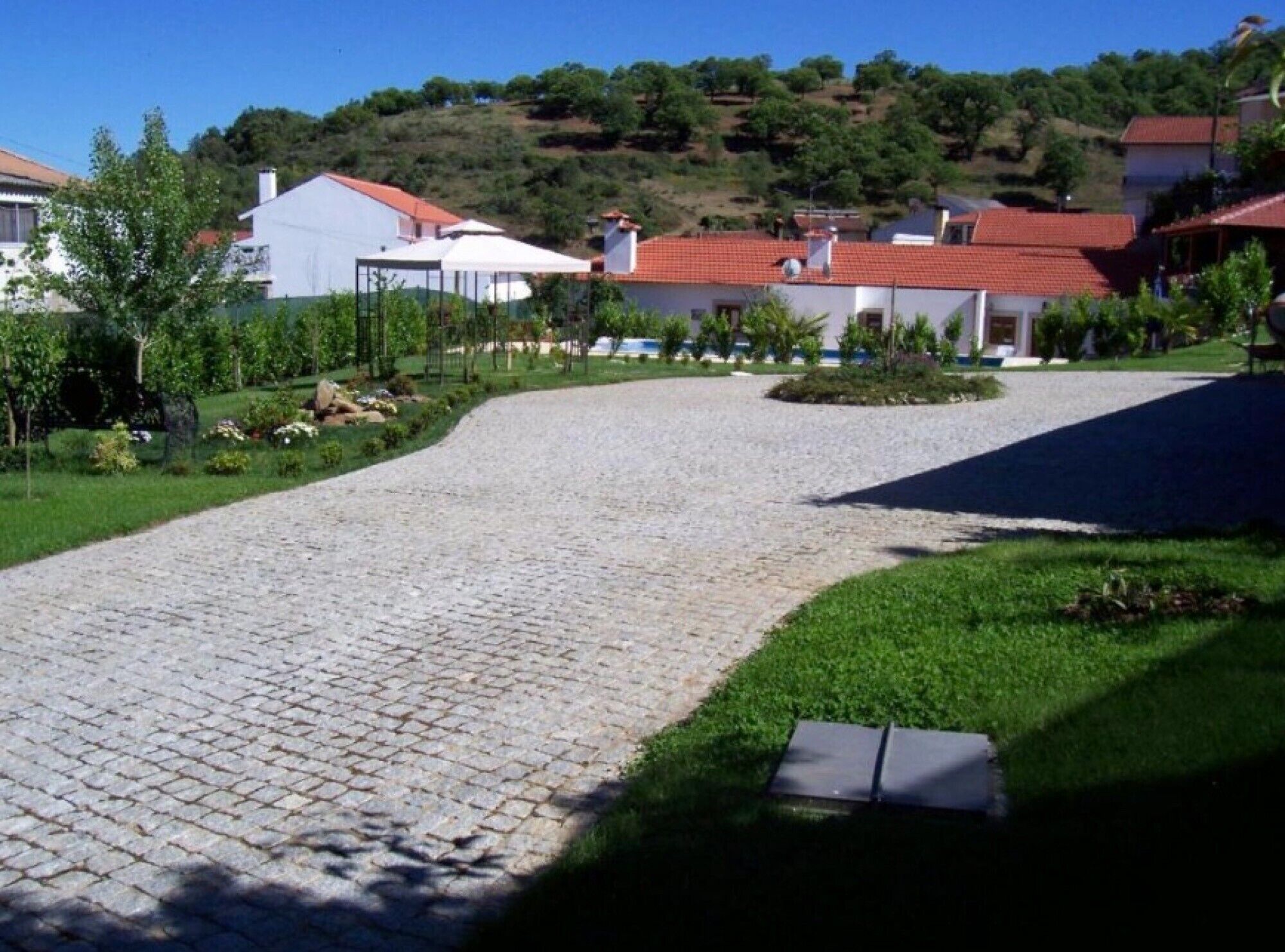 A cobblestone driveway leading to a residential area with houses, greenery, and a hilly backdrop.