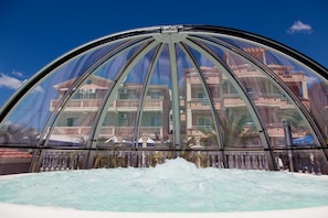 A large, arched glass enclosure with a pool in the foreground and a building with balconies in the background.