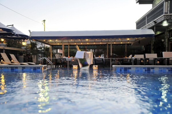 A swimming pool with lounge chairs and umbrellas, a covered seating area, and a building with a balcony.
