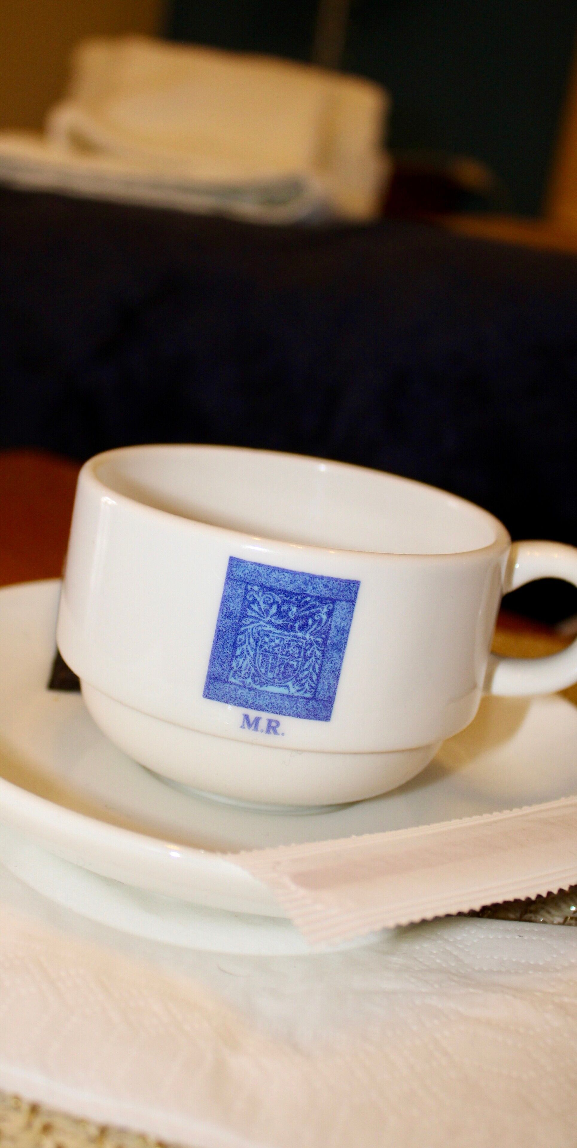 Una taza blanca con etiqueta azul sobre un plato, colocada sobre una mesa con una servilleta debajo.