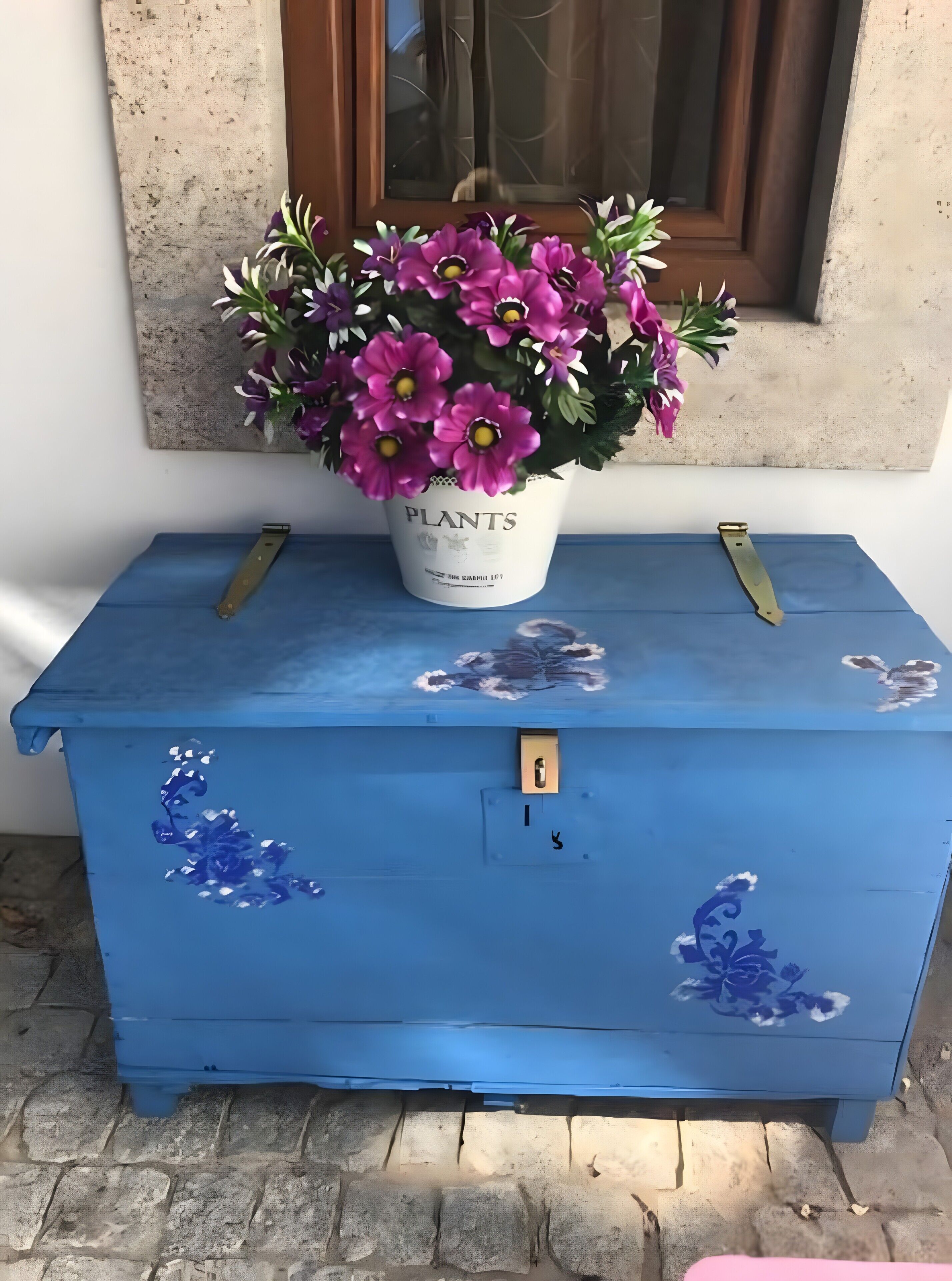 A blue chest of drawers with floral decorations, a potted plant with purple flowers, and a window in the background.