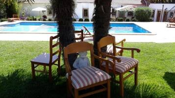 A poolside area with two wooden chairs and a table, surrounded by grass and palm trees.