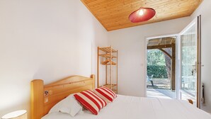 Een slaapkamer met een houten hoofdbord, een bed met wit beddengoed en gestreepte kussens, een bedlampje en uitzicht op het buitengebied door een glazen deur.