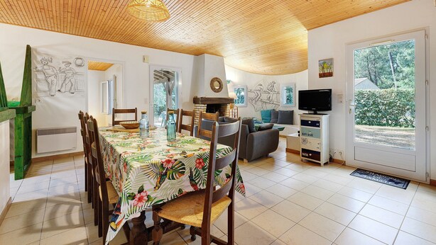 Een eetgedeelte met een houten plafond, een tafel met een bloemrijk tafelkleed, houten stoelen, een bank, een televisie en uitzicht op groen buiten.