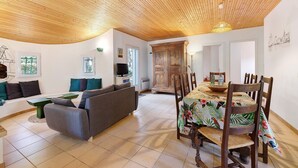 Een woonkamer met een bank, een eettafel met stoelen, een houten plafond en uitzicht op buiten.