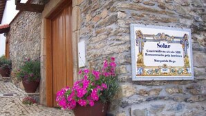 Un edificio de piedra con una puerta de madera, un cartel que indica que fue construido en el siglo XIII, y una maceta con flores rosadas.