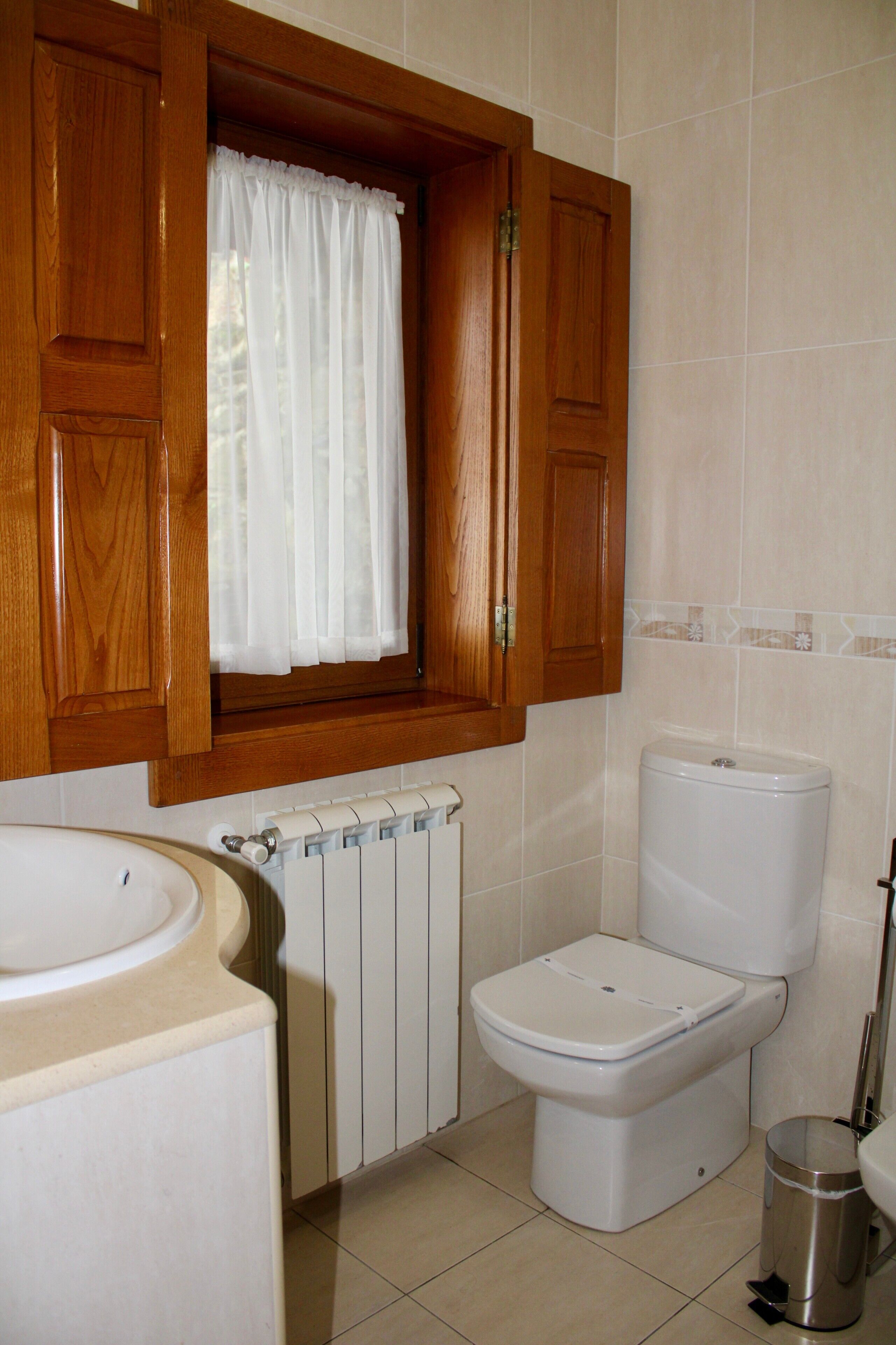 A bathroom with wooden cabinets, a white toilet, a radiator, and a window with a white curtain.