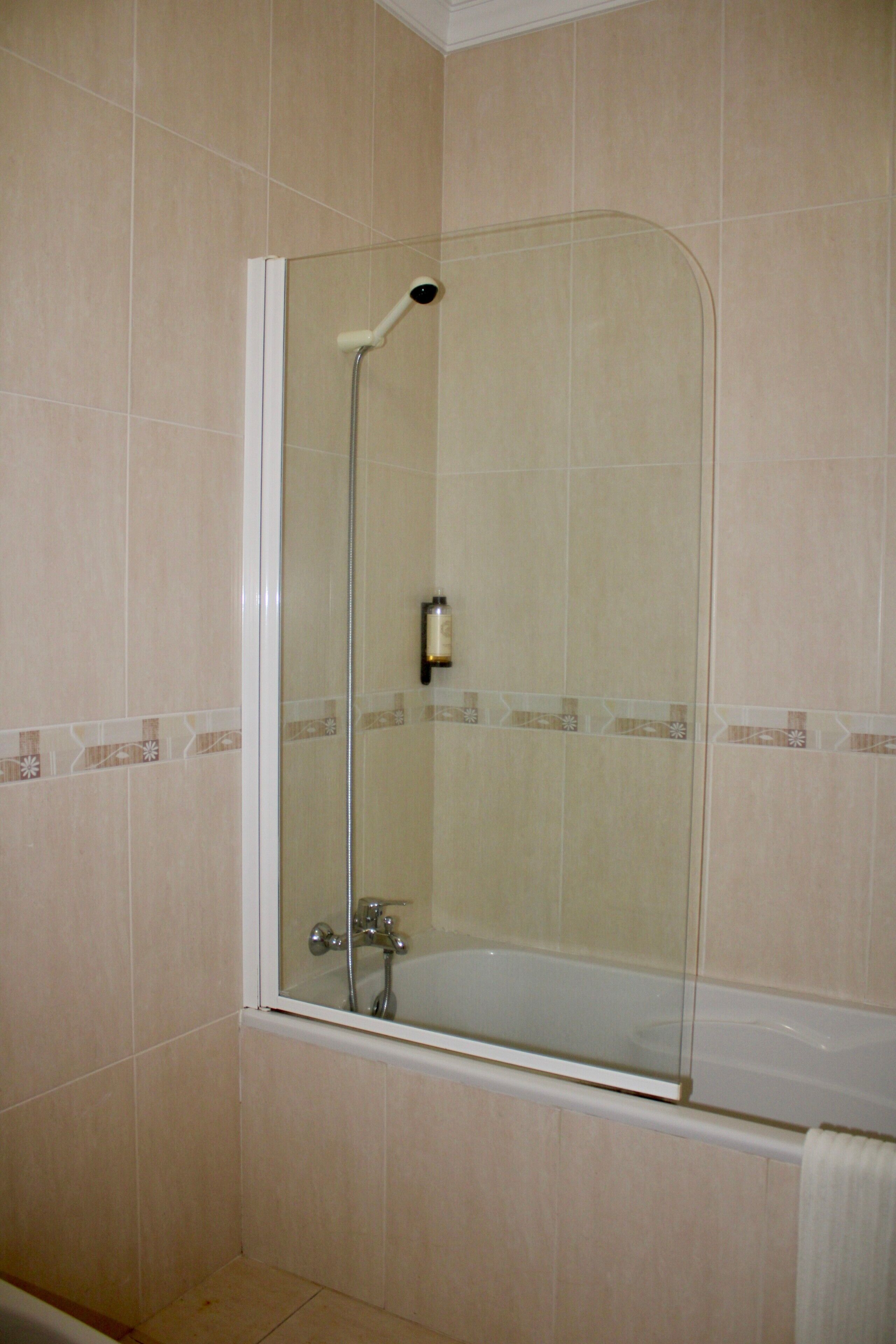 A glass-enclosed shower with a handheld showerhead, a handheld faucet, and a white bathtub with a towel.