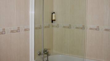 A glass-enclosed shower with a handheld showerhead, a handheld faucet, and a white bathtub with a towel.