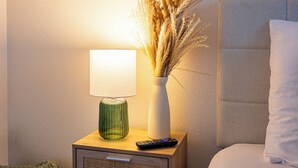 Una mesita de noche con una lámpara, un florero con plantas secas, un control remoto y una cama con un almohadón.