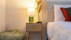 Una cama bien hecha con almohadas blancas y rojas, una lámpara de noche y una mesita de noche con un florero de plantas secas.
