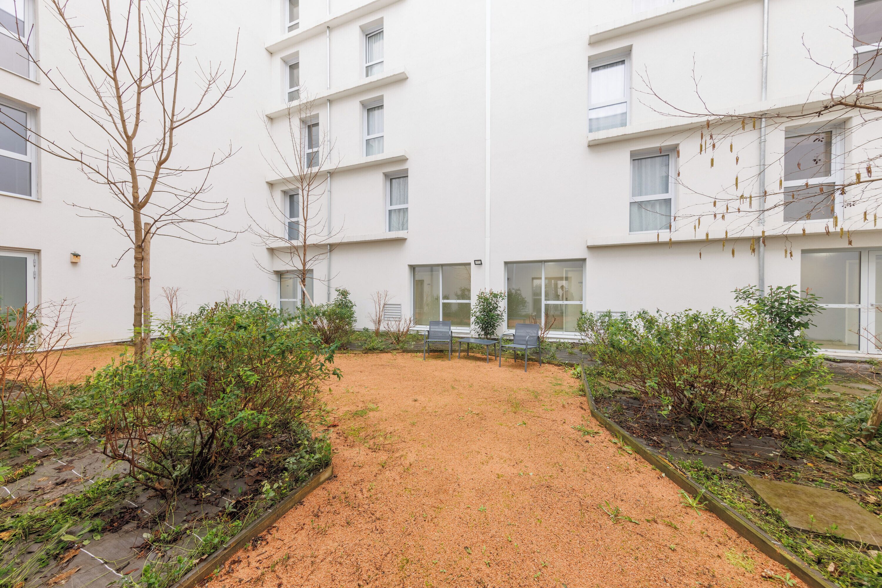 A courtyard with a sandy ground, two rows of chairs, and a building with multiple windows and balconies.
