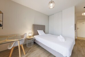 Ein modernes Hotelzimmer mit einem Glastisch, einem Einzelbett mit weißer Bettwäsche, einem runden Beistelltisch mit Lampe und Holzboden.
