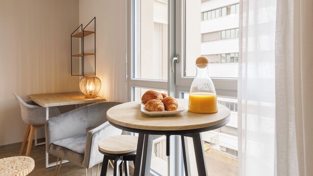 A modern room with a round table, a chair, a lamp, a plate with croissants, and a glass of orange juice. - 6 Le Loki - Tramway - Wifi (Clermont-Ferrand)