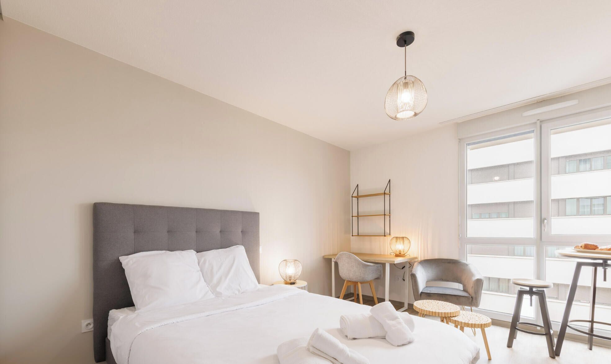 Ein Hotelzimmer mit Bett, Schreibtisch, Stuhl, kleinem Tisch und einem Balkon mit Blick auf Gebäude.