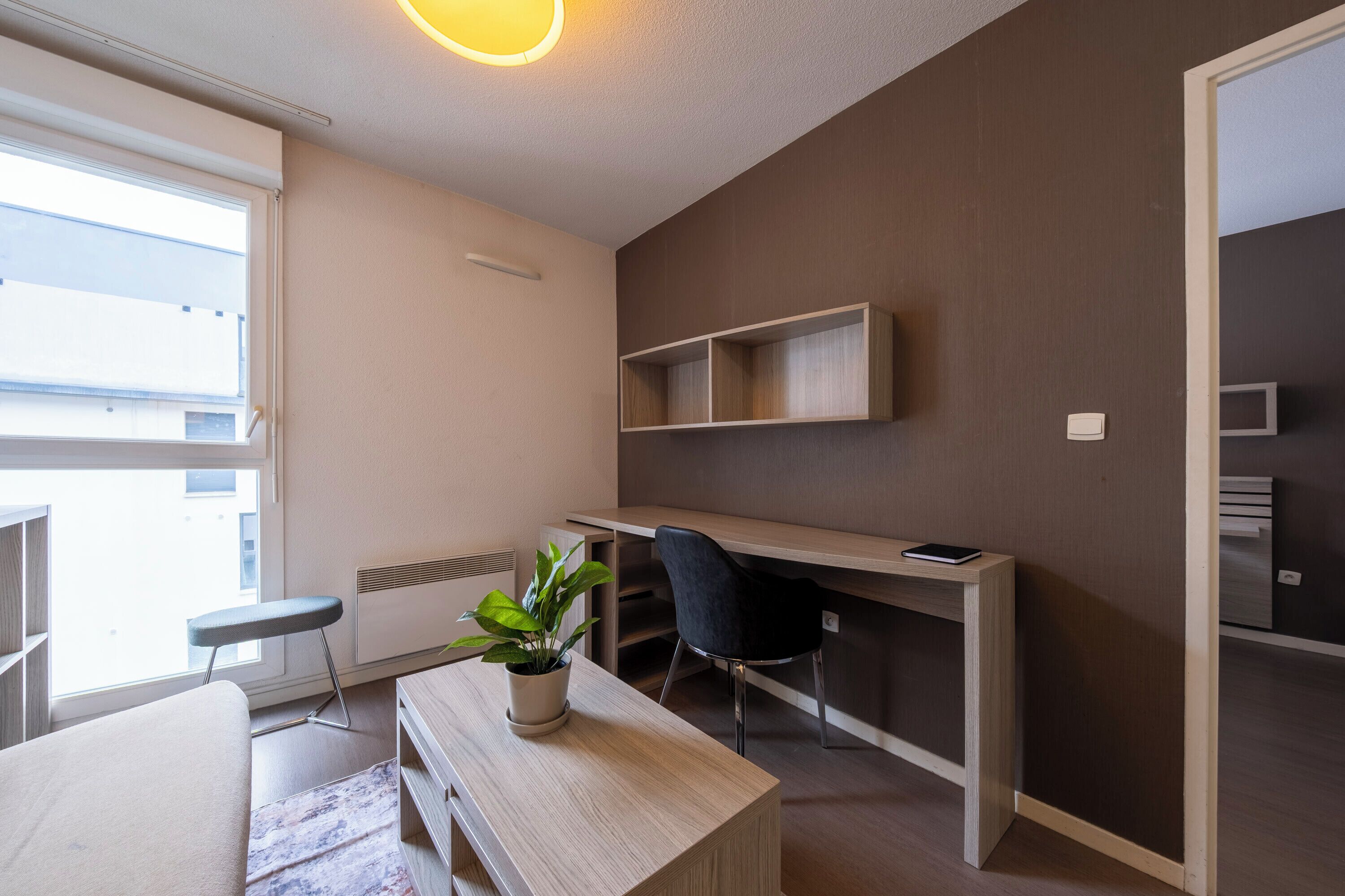 Habitación moderna y bien iluminada con un escritorio de madera, una silla, un pequeño banco y una planta en maceta.