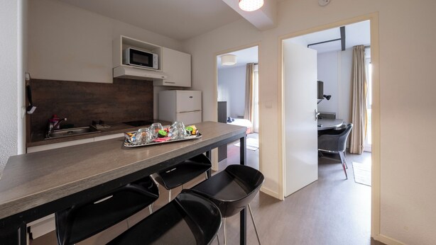 A modern kitchen with a microwave, sink, and bar stools. There is a dining table with a tray of glasses and a bottle. The room has a neutral color scheme and a ceiling light. - 8 - L'Envol - Gare - Wifi (Toulouse)