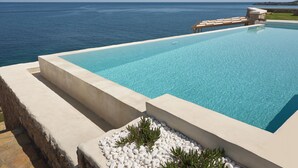 Een modern overloopzwembad met helderblauw water, omringd door stenen muren en witte kiezelstenen, met uitzicht op een rustige zee.