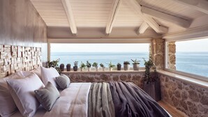Een slaapkamer met een bed, een stenen muur, een raam met uitzicht op zee en verschillende potplanten op de vensterbank.