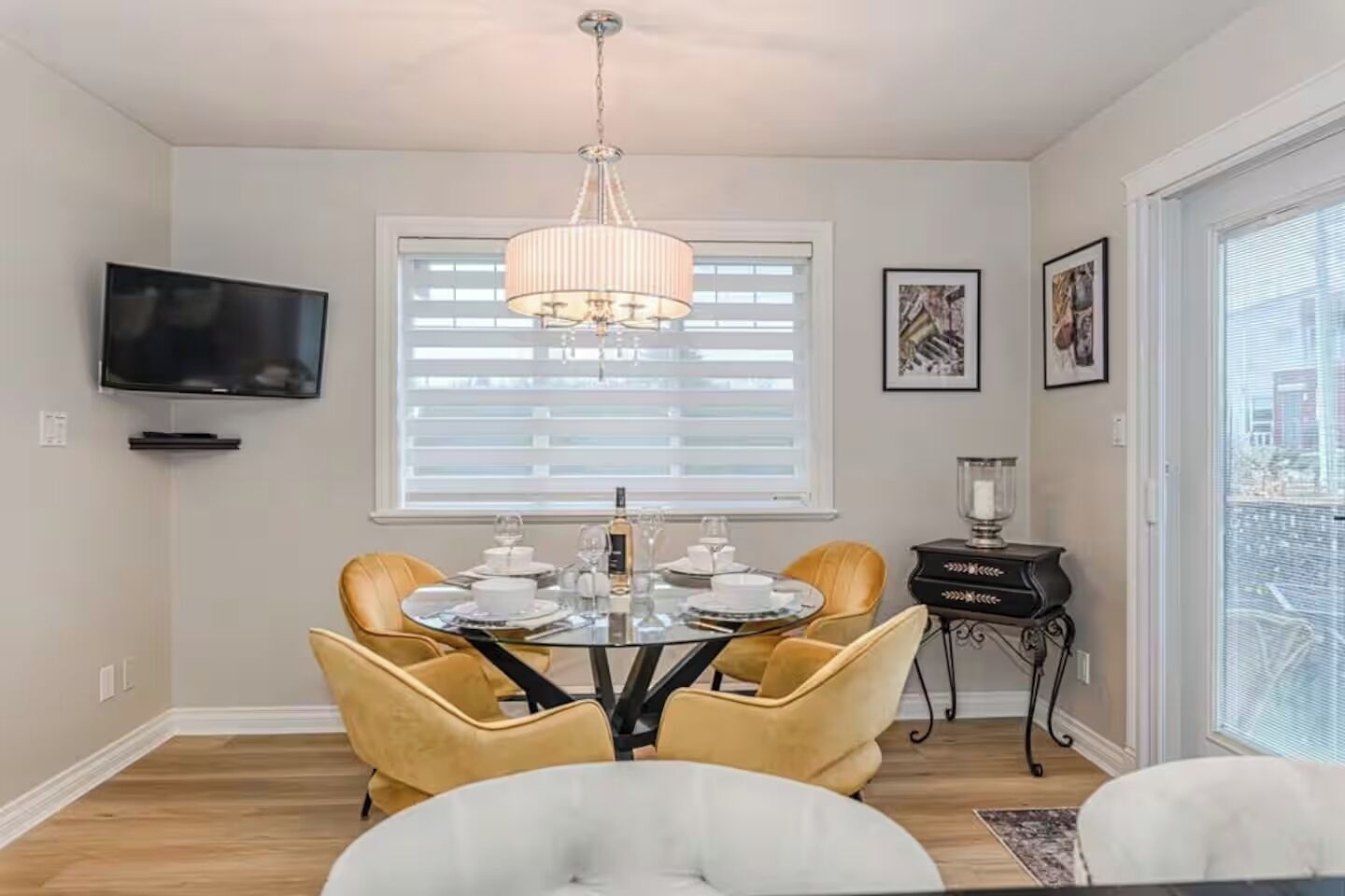 Un comedor con una mesa redonda, cuatro sillas, una lámpara de araña, un televisor montado en la pared y una ventana con persianas.