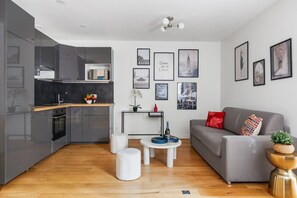 A modern kitchen with grey cabinets, a wooden countertop, and a built-in oven. There is a dining area with a white table and two stools. The living room features a grey sofa with red and patterned cushions. - ClickYourFlat 256 - Cosy Apt - Arc de Triomphe (Paris)