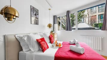 Une chambre d’hôtel moderne avec un lit, une couverture rouge, des oreillers blancs, un lustre doré et une vue sur les bâtiments par la fenêtre.