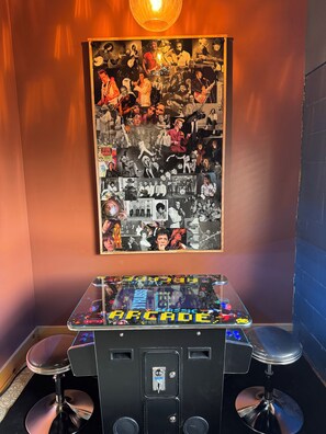 A room with a wall-mounted collage of black and white photos, a glass table with arcade game graphics, and two bar stools.