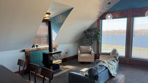 A cozy loft bedroom with a sloped ceiling, a fireplace, a dining area, and large windows with blinds. - Cozy Gem With Stunning Mississippi River View (Albany)