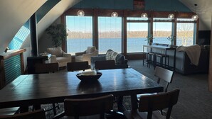 A dining area with a large table, chairs, and a view of a body of water through large windows. - Cozy Gem With Stunning Mississippi River View (Albany)
