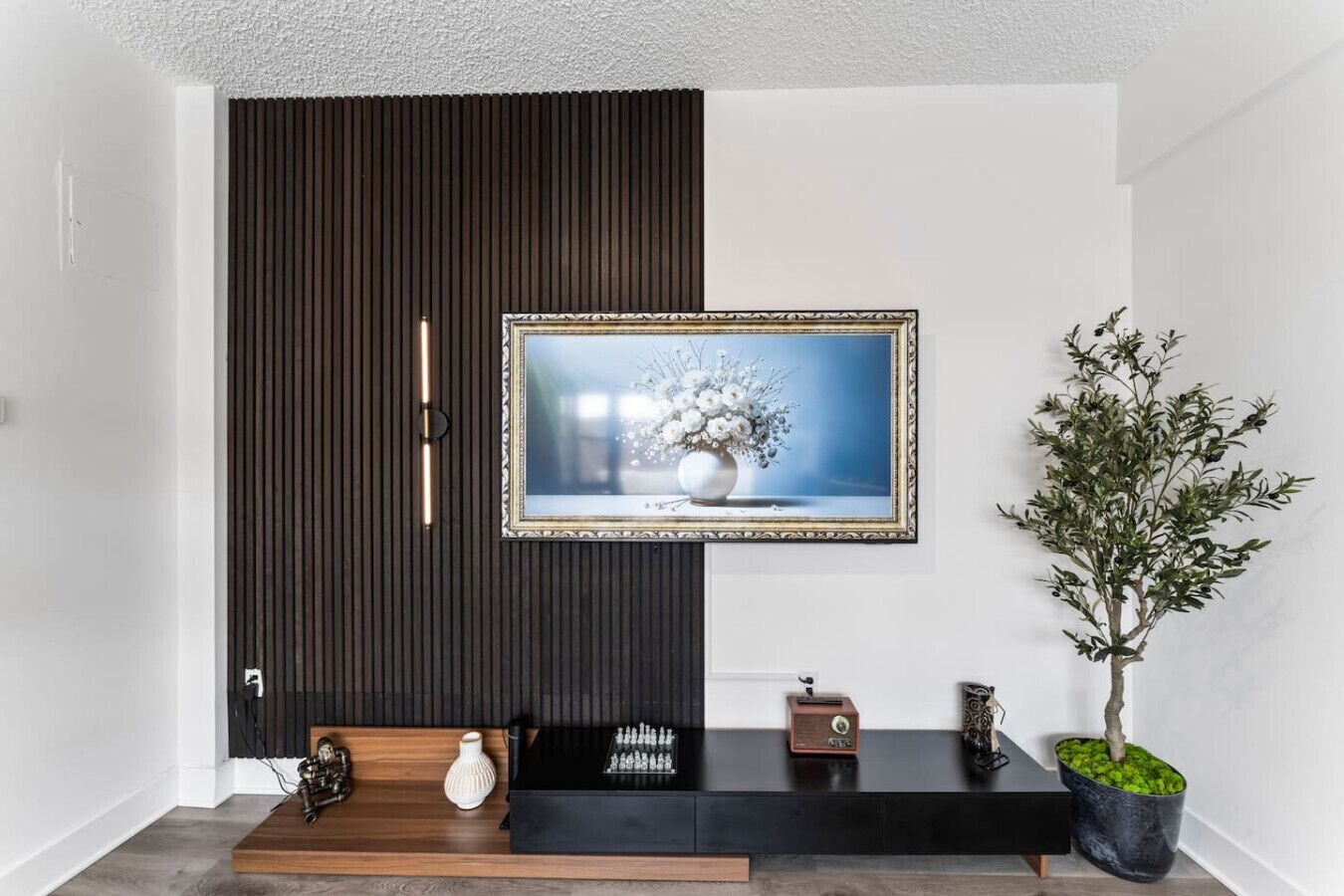 A modern living room with a dark wooden panel wall, a framed picture of flowers, a chessboard, a vintage radio, and a potted plant.