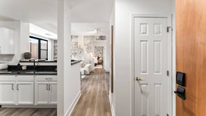 A modern kitchen with white cabinets, a black countertop, and a window. A hallway leads to a bedroom with a wooden door and a bedside table. - Solara – Luxury 1BR Downtown (Stamford)