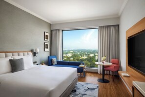 Habitación de hotel con una cama grande, un sofá azul, una mesita, una silla roja y un ventanal con vistas a la ciudad.