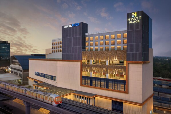 Un hotel moderno con un cartel llamativo de 'Bansal Plaza', un letrero de 'Hyatt Place' y vistas a las vías del tren de abajo.