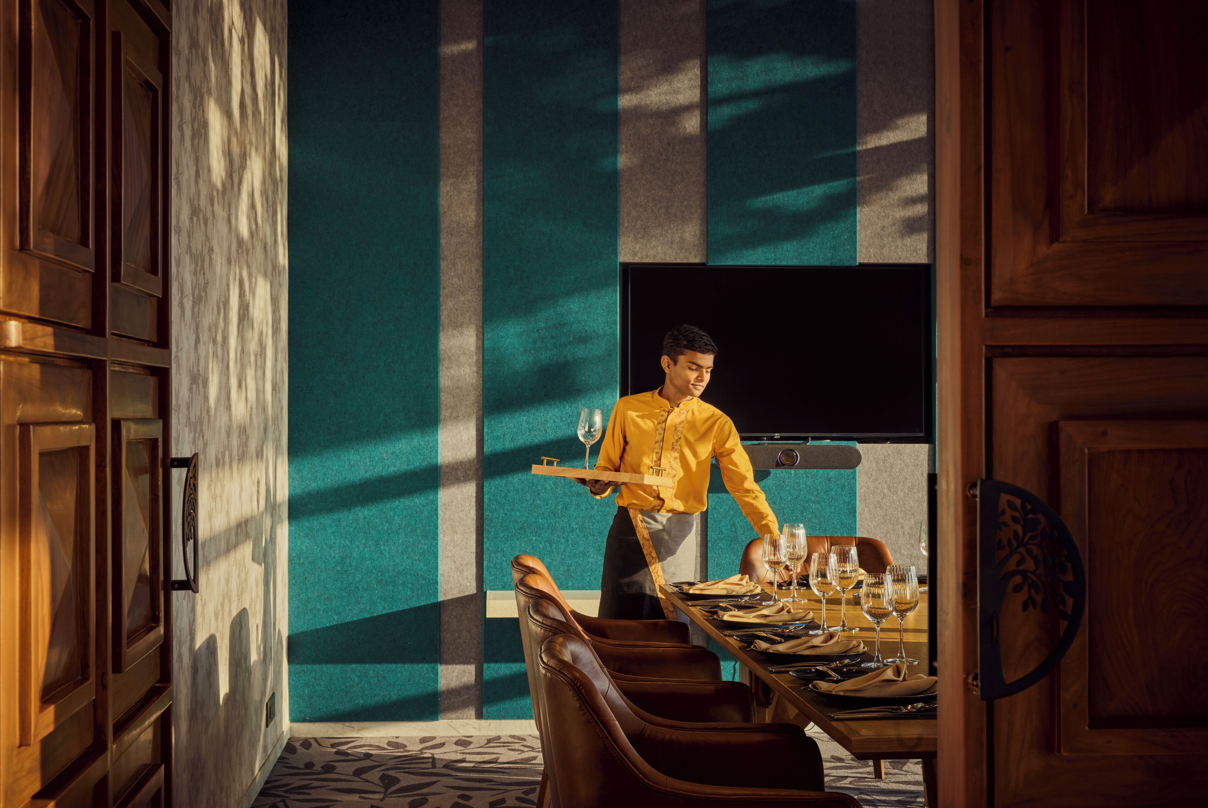 A waiter carrying a tray with wine glasses and a bottle, standing in front of a table set for dining.