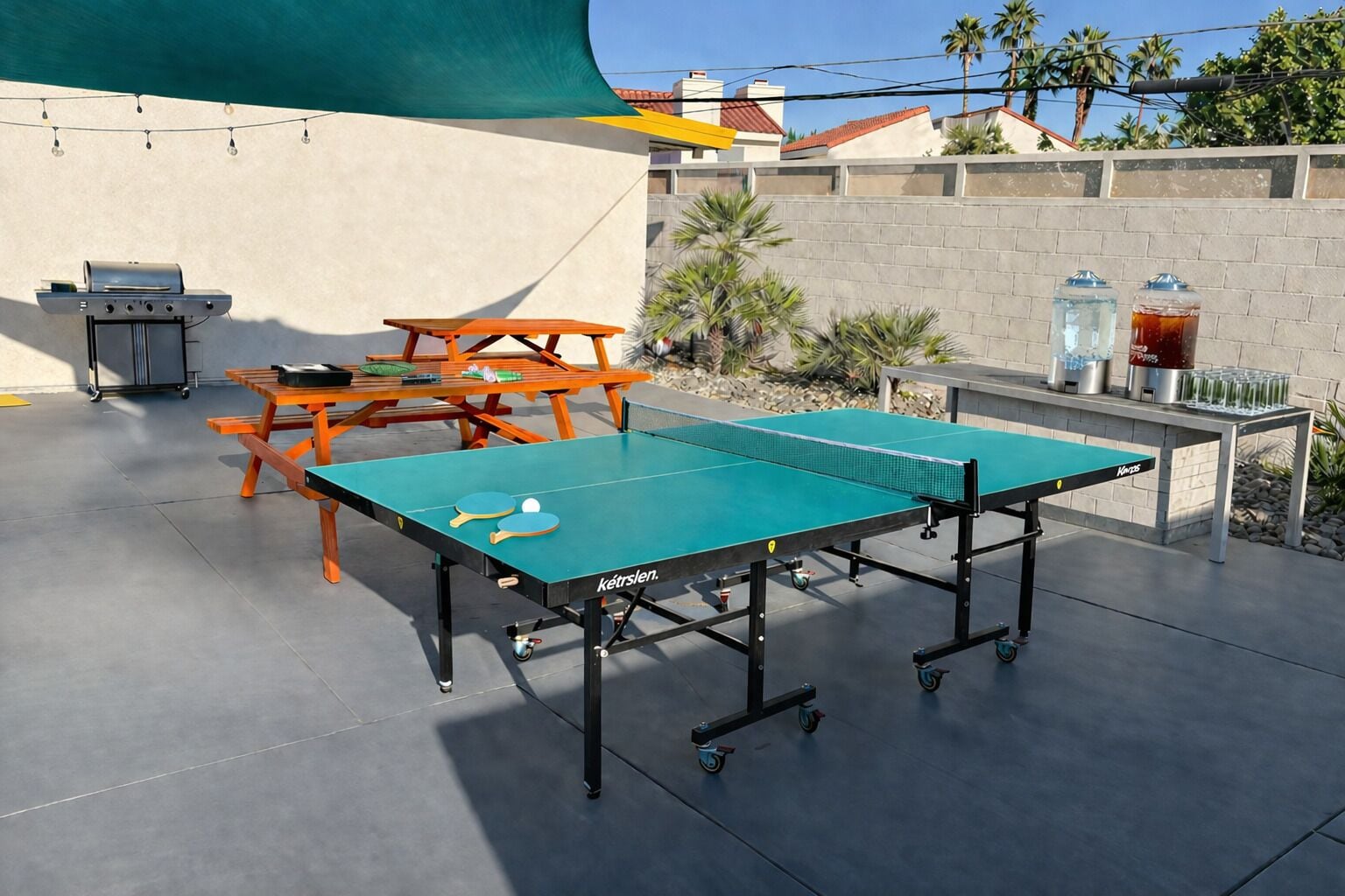 A ping pong table with a net, a grill, and a table with drinks on a patio.