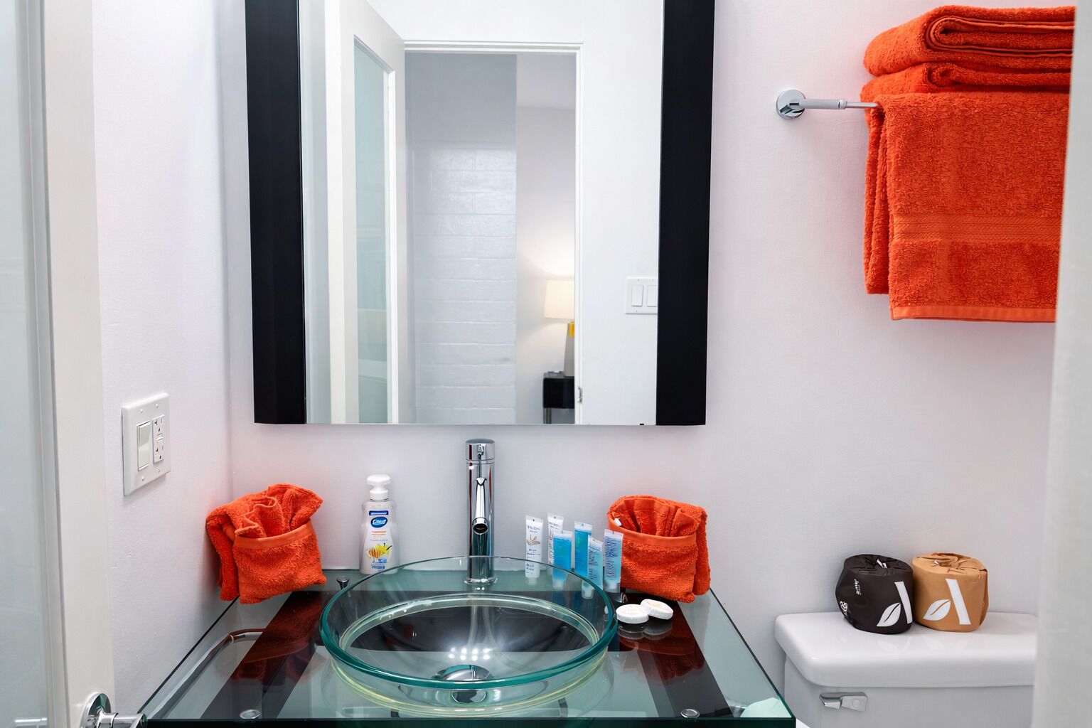 A bathroom with a glass sink, a mirror, a toilet, and orange towels.