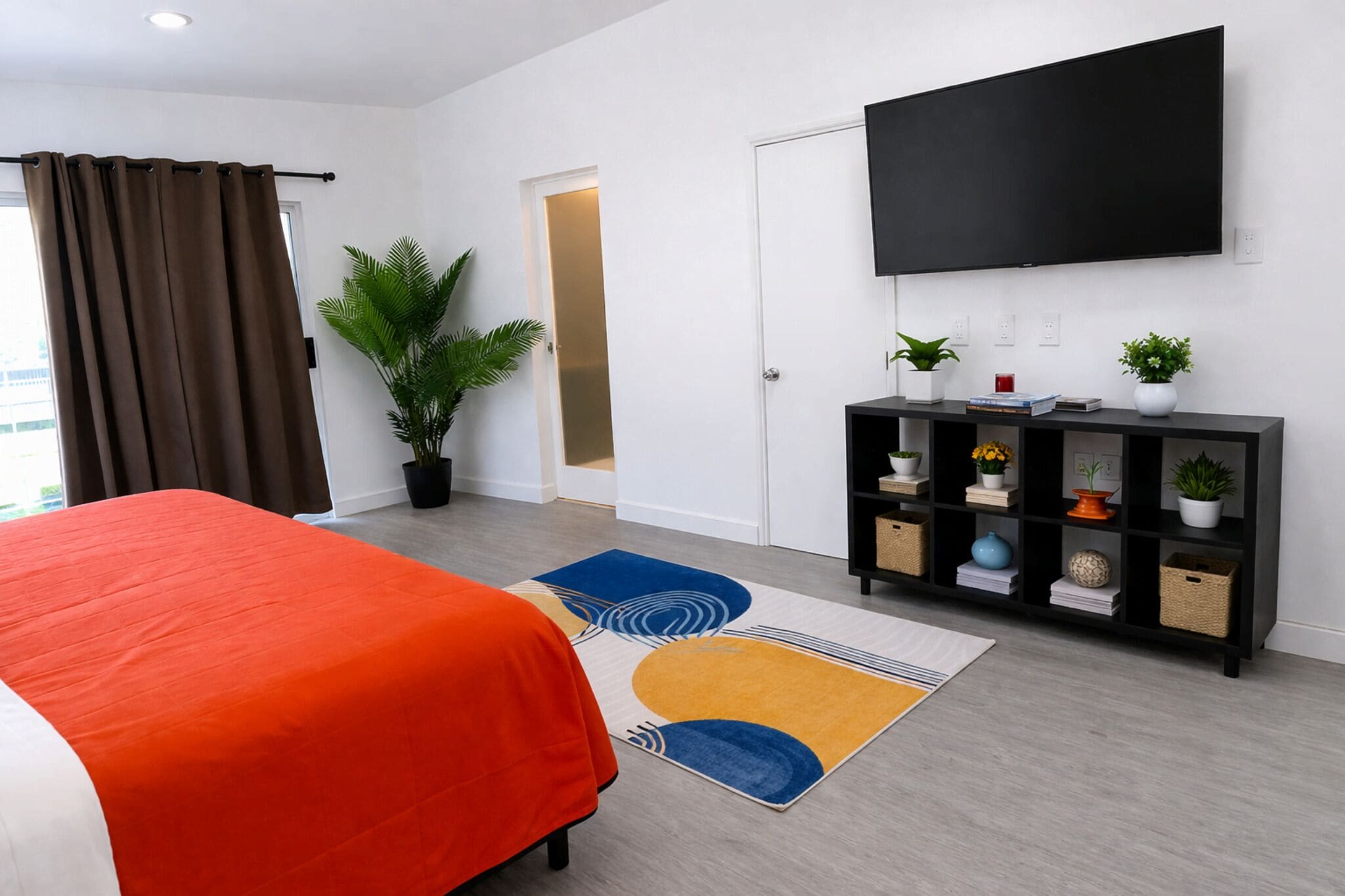 A modern hotel room with a bed, a TV, a bookshelf, and a potted plant.