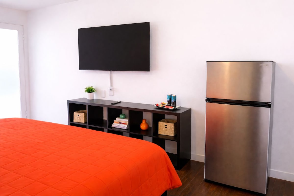Habitación de hotel con cama, televisor de pantalla plana montado en la pared, refrigerador y una mesa consola con varios objetos.