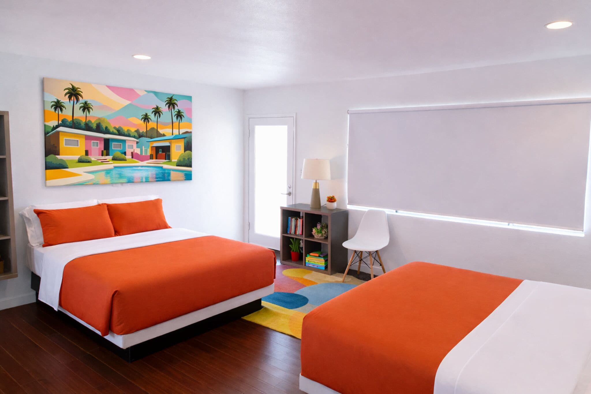 A hotel room with two beds, a colorful painting, a bookshelf, and a window with blinds.