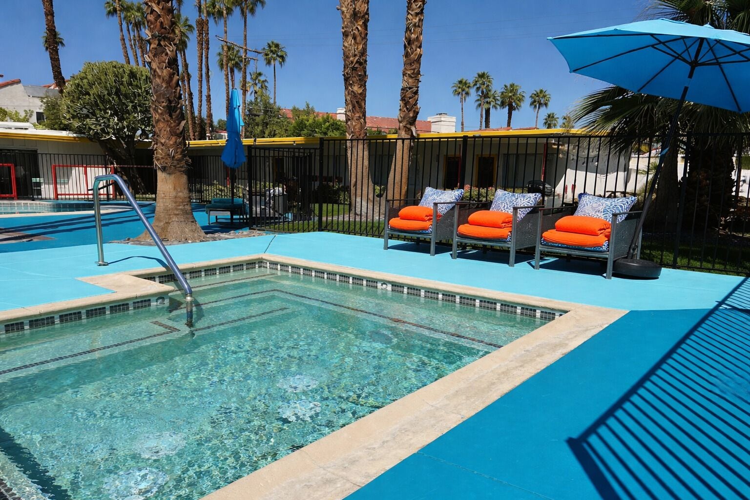 A swimming pool with lounge chairs and umbrellas, surrounded by palm trees and a fence.