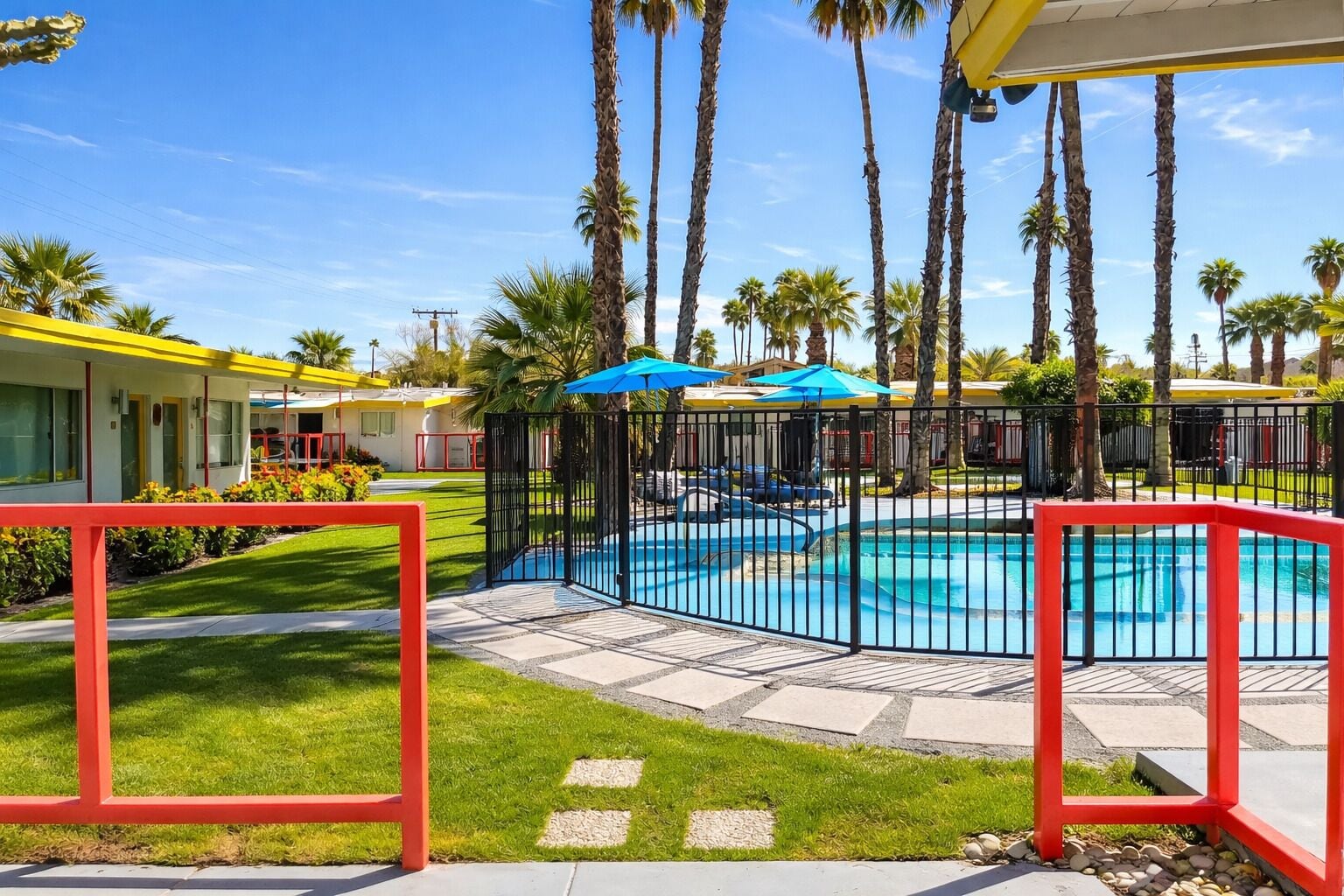 A pool area with a fenced pool, palm trees, and a building with yellow and red accents.